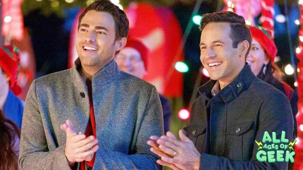 Jonathan Bennett and another Hallmark star at a festive holiday event, surrounded by Christmas lights and decorations, looking cheerful and engaged in the festive atmosphere. The All Ages of Geek logo is visible in the corner of the image.