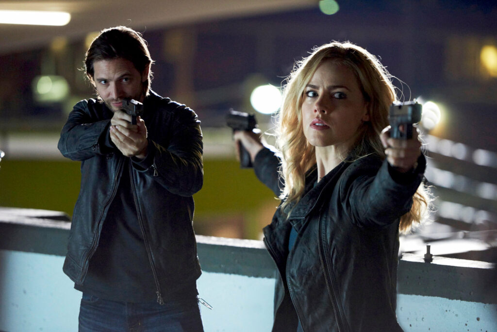 A dramatic scene from the TV series '12 Monkeys,' featuring the characters James Cole and Dr. Cassandra Railly. Both characters are aiming handguns, with intense and focused expressions. James Cole, portrayed by Aaron Stanford, is on the left wearing a black leather jacket. Dr. Cassandra Railly, portrayed by Amanda Schull, is on the right, also in a black leather jacket. The background shows an urban setting at night, with blurred lights adding to the tension of the scene. One of the iconic New Jersey Film Locations.