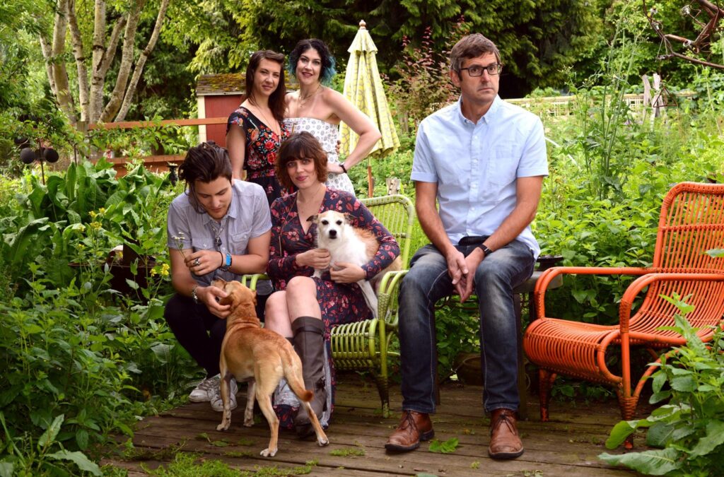 A group photo of the cast from 'Altered States.' They are outdoors in a lush garden setting. Three women are standing in the back, smiling and posing, while two people are seated in front. The woman seated on the left is holding a small dog, and a man in a light blue shirt and glasses is seated on the right, looking off to the side. Another man kneels next to a small brown dog, engaging with it. The scene is casual and relaxed, with greenery and a wooden deck in the background. One of the iconic New Jersey Film Locations.