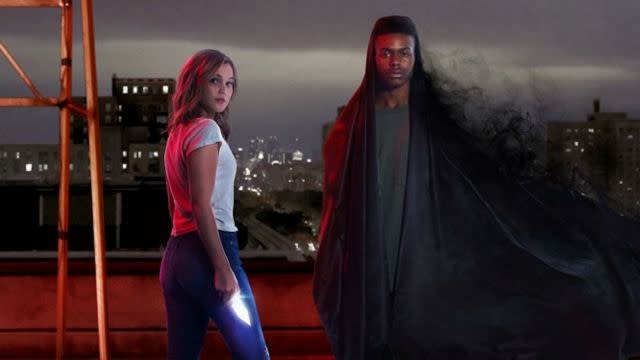 A scene featuring the characters Cloak and Dagger from the Marvel series 'Cloak & Dagger.' Dagger, on the left, is a young woman with blonde hair, wearing a white t-shirt and blue jeans, holding a glowing light dagger. Cloak, on the right, is a young man with dark skin, partially shrouded in a dark, flowing cloak that blends into the night. They stand on a rooftop with a cityscape in the background under a cloudy night sky, giving the scene a mysterious and dramatic atmosphere.