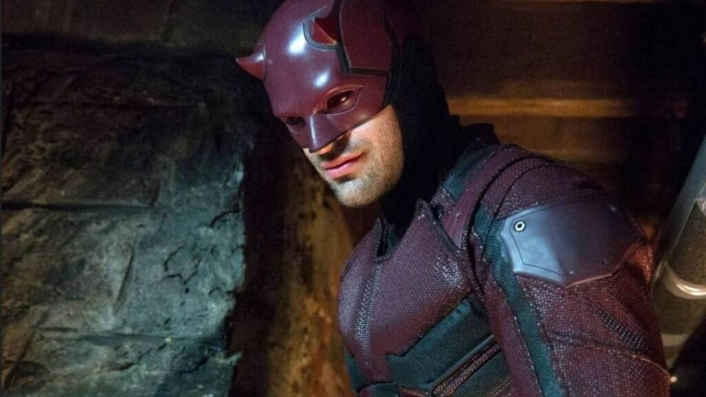 A scene from the Marvel series 'Daredevil,' featuring the character Daredevil in his red suit and mask with small horns. The character, portrayed by Charlie Cox, is shown in a dimly lit environment, possibly a dark alley or underground setting. He is looking to the side with a serious expression, highlighting the intense and gritty atmosphere of the show.