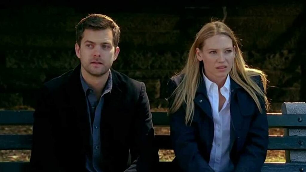 A scene from the TV series 'Fringe,' featuring the characters Peter Bishop and Olivia Dunham. Peter, portrayed by Joshua Jackson, is on the left, wearing a dark jacket and looking off to the side. Olivia, portrayed by Anna Torv, is on the right, wearing a dark coat over a white shirt, also looking to the side with a serious expression. They are sitting on a park bench, with a dark, blurred background, suggesting an intense or contemplative moment. One of the iconic New Jersey Film Locations.