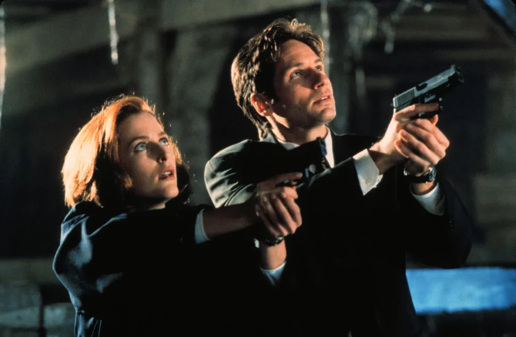 A scene from 'The X-Files' featuring FBI agents Dana Scully, played by Gillian Anderson, and Fox Mulder, played by David Duchovny. Both characters are standing close together, looking upwards with serious expressions. Scully is on the left, holding a flashlight, while Mulder is on the right, holding a handgun. The background is dark and mysterious, adding to the show's suspenseful atmosphere. One of the iconic New Jersey Film Locations.