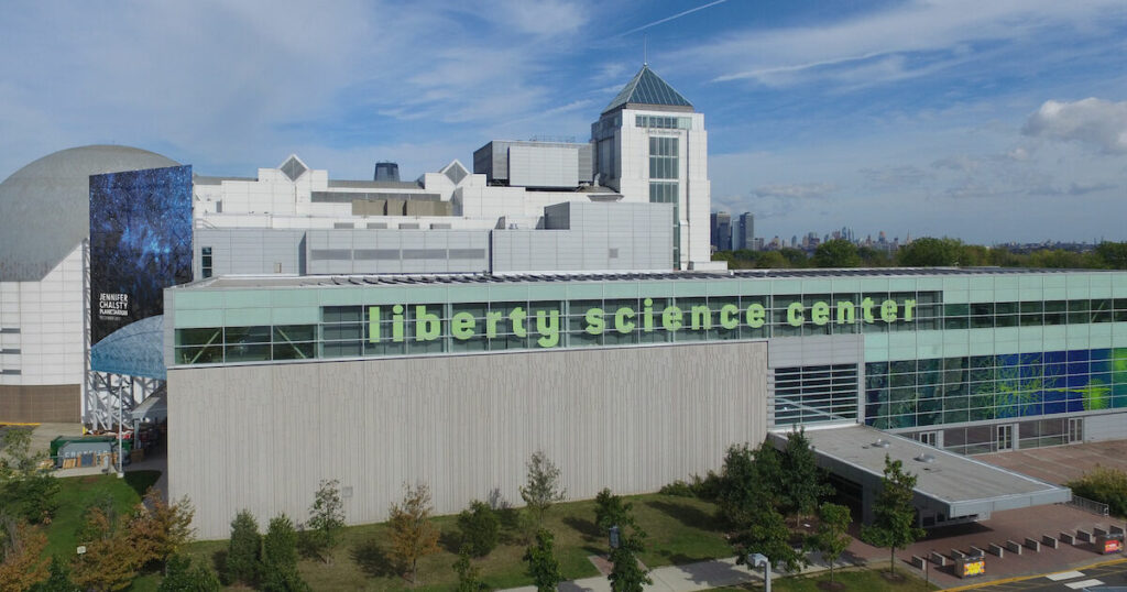 A daytime view of the Liberty Science Center in Jersey City, New Jersey. The modern building features a large, prominent sign with green letters spelling 'liberty science center.' The structure includes a mix of glass and concrete elements, with a dome and various geometric shapes adding to its architectural interest. The background includes a view of the New York City skyline under a partly cloudy sky. One of the iconic New Jersey Film Locations.