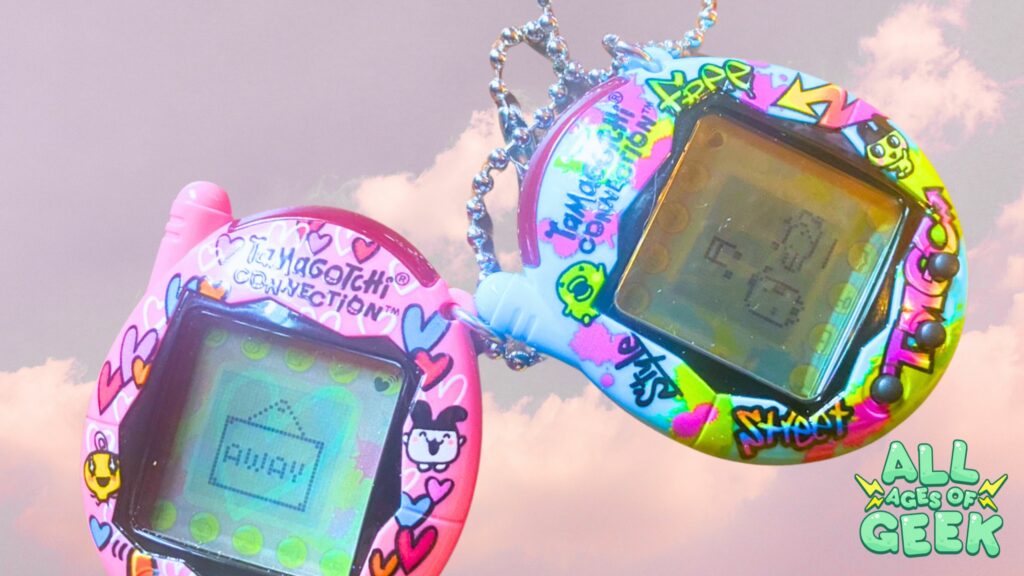 wo Tamagotchi Connection virtual pets, one with a pink shell featuring heart designs and another with a colorful graffiti-style shell, are shown against a pastel sky background. Both devices display pixelated characters on their screens. The All Ages of Geek logo is visible in the bottom right corner.