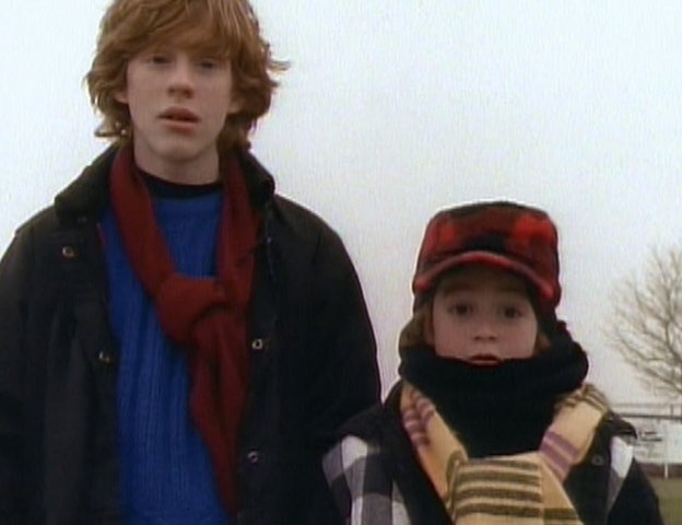 A scene from 'The Adventures of Pete & Pete' featuring the two main characters, Big Pete and Little Pete, standing side by side. Big Pete, on the left, has red hair and is wearing a blue sweater, black jacket, and red scarf. Little Pete, on the right, is bundled up in a plaid jacket, yellow scarf, and red and black checkered hat. The background is outdoors, with a tree and overcast sky visible.