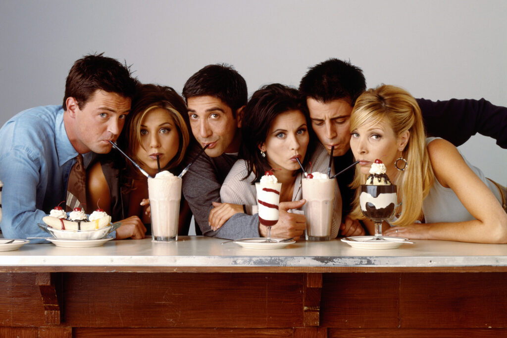 In the image, six friends from the iconic 90s sitcom "Friends" are gathered around a counter, each enjoying a different ice cream or milkshake treat. From left to right, you can see Chandler, Rachel, Ross, Monica, Joey, and Phoebe. They're all sipping from their straws, looking directly at the camera with playful and amused expressions. The image captures the fun and camaraderie that defined the show, making it a beloved favorite among fans.