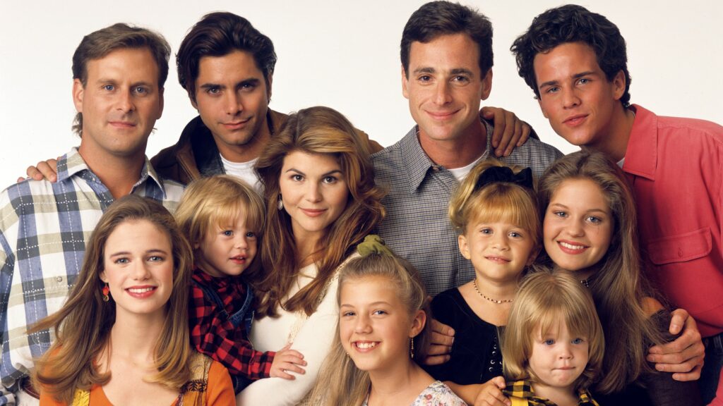 In the image, the cast of the beloved 90s sitcom "Full House" is gathered together, showcasing the warmth and closeness of the Tanner family. In the back row from left to right are Joey Gladstone, Jesse Katsopolis, Danny Tanner, and Steve Hale. In the middle row are Becky Katsopolis and D.J. Tanner, holding Michelle Tanner. In the front row are Stephanie Tanner, Michelle Tanner, and D.J. Tanner. The family is smiling warmly, exuding a sense of unity and love that made the show a favorite among viewers. Their casual and comfortable attire adds to the cozy, family-friendly vibe of the series.