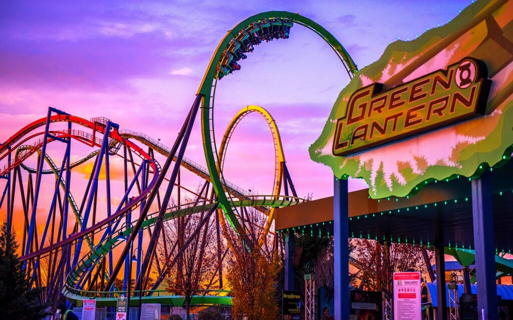 Photograph of the Green Lantern roller coaster at Six Flags Great Adventure during sunset. The vibrant sky is filled with shades of pink, purple, and orange, creating a dramatic backdrop for the looping tracks of the roller coaster. The roller coaster's structure is painted in bright colors, including green, red, and blue, and the sign for 'Green Lantern' is prominently displayed in the foreground with a green and yellow color scheme. The image captures the excitement and energy of the amusement park ride against a picturesque evening sky. Image used for Pokemon Go New Jersey.