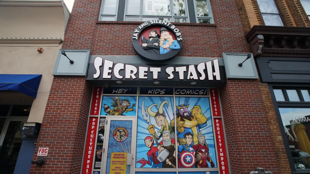 Exterior of Jay and Silent Bob's Secret Stash, a comic book store in Red Bank, New Jersey. The storefront features a colorful mural of comic book characters like Loki, Thor, and Iron Man, along with a large sign displaying the store's name above the entrance. The store has a classic brick exterior, and a circular logo above the sign depicts cartoon versions of Jay and Silent Bob. Example of Central Jersey does exist. 