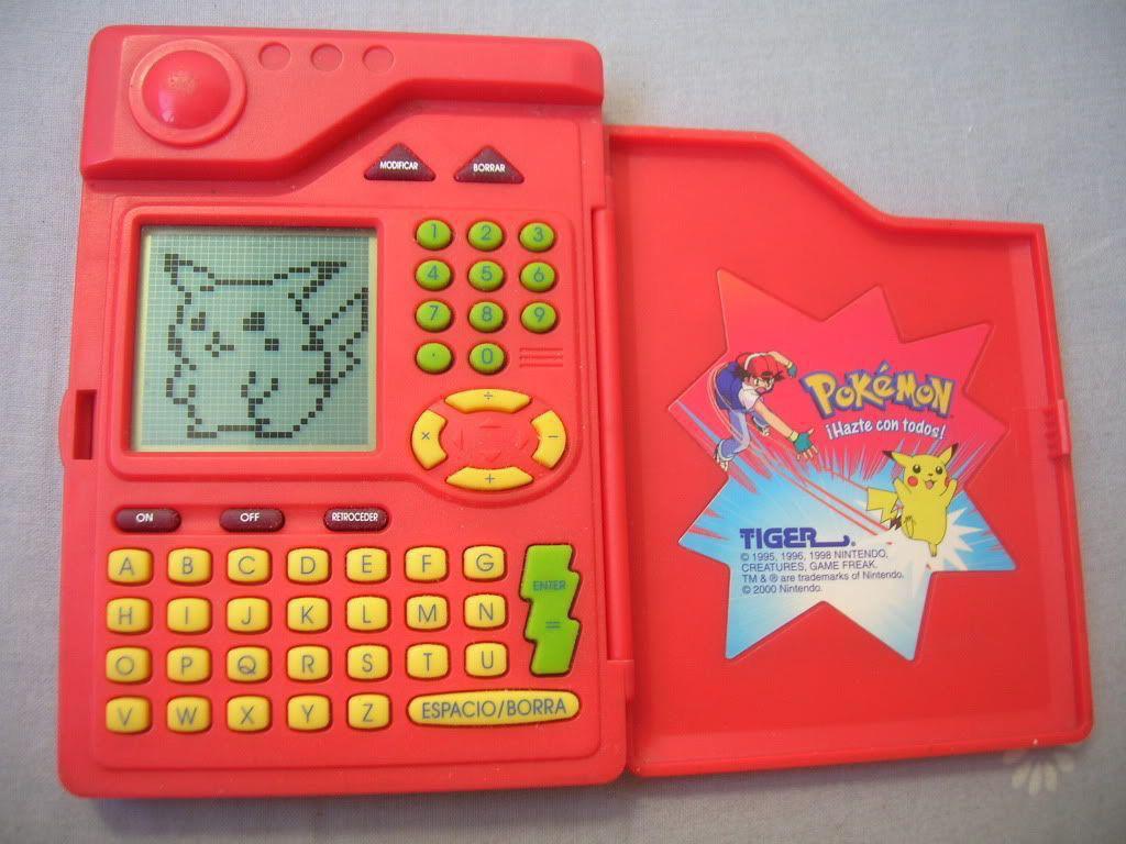 Image of a vintage Pokémon Pokédex toy from the 1990s, produced by Tiger Electronics. The device is red and designed to resemble the iconic Pokédex from the Pokémon series. The left side features a small monochrome screen displaying a pixelated image of Pikachu, with a full QWERTY keyboard below it, along with buttons for navigation, on/off, and other functions. The right side has a plastic cover with a sticker showing Ash Ketchum and Pikachu, along with the Pokémon logo and the phrase '¡Hazte con todos!' (Spanish for 'Gotta catch 'em all!'). The toy has a nostalgic design, capturing the look and feel of the original Pokédex from the Pokémon anime.