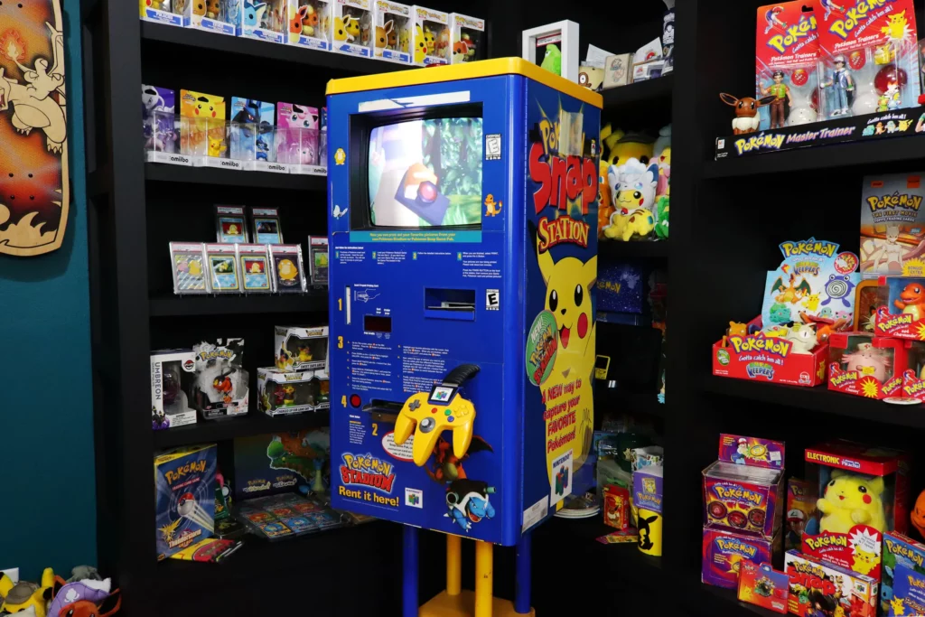 Photograph of a vintage Pokémon Snap Station kiosk, originally found in Blockbuster stores, designed for players to print out photos taken in the Pokémon Snap game on the Nintendo 64. The kiosk is blue and yellow, featuring Pikachu on the side with the 'Pokémon Snap Station' logo. The front of the kiosk has a screen displaying gameplay and a Nintendo 64 controller attached for interaction. The kiosk is surrounded by shelves filled with various Pokémon merchandise, including action figures, trading cards, and plush toys, creating a nostalgic atmosphere for fans of the franchise.