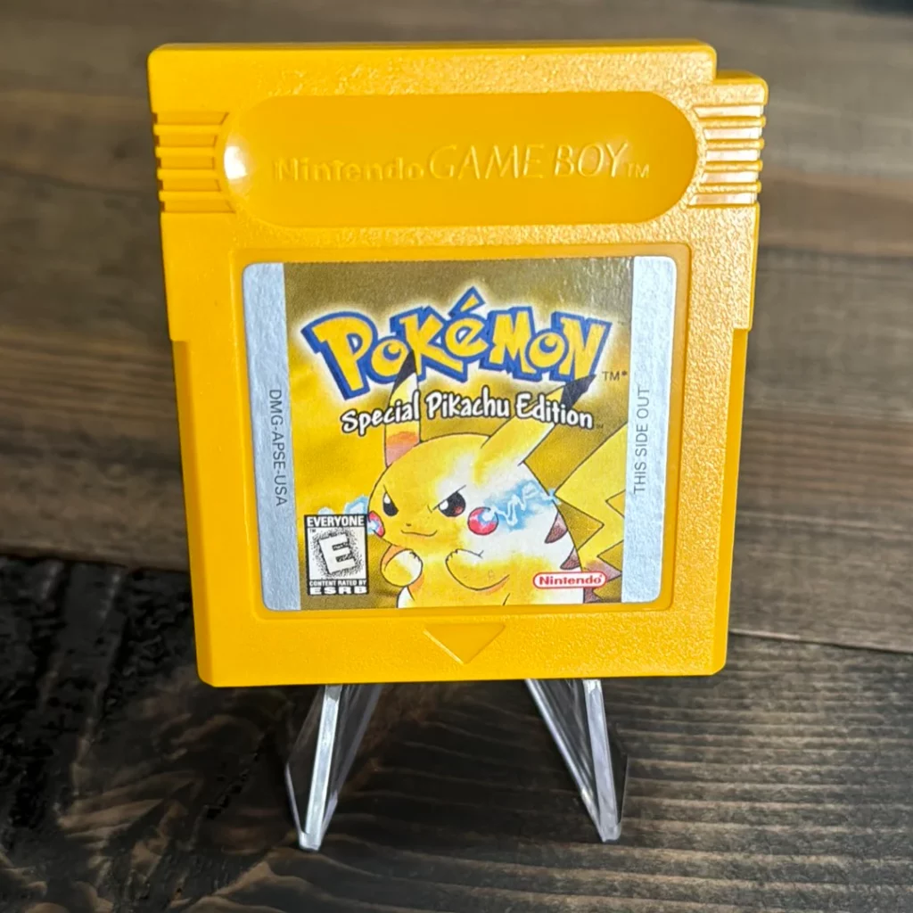 Close-up image of a yellow Game Boy cartridge for the game Pokémon: Special Pikachu Edition, commonly known as Pokémon Yellow. The cartridge is displayed on a small clear stand against a wooden surface. The label on the cartridge features an illustration of Pikachu with an intense expression, with the game title above it. The text 'Nintendo GAME BOY' is embossed at the top of the cartridge, and the ESRB rating is marked as 'Everyone.' The cartridge has a distinct bright yellow color, matching the theme of the game.