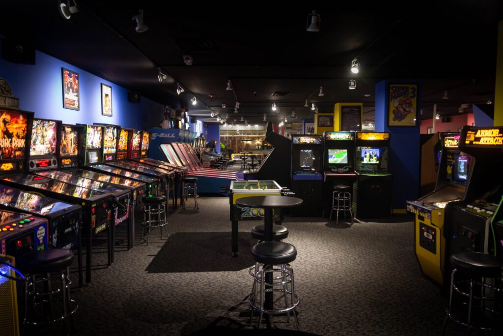 Interior of Yestercades, a retro arcade in Westfield, New Jersey. The arcade features rows of vintage pinball machines and classic arcade games like Pac-Man and Armor Attack. The room is dimly lit, with small tables and stools set up between the machines, creating a nostalgic atmosphere for gaming enthusiasts. Example of Central Jersey does exist. 