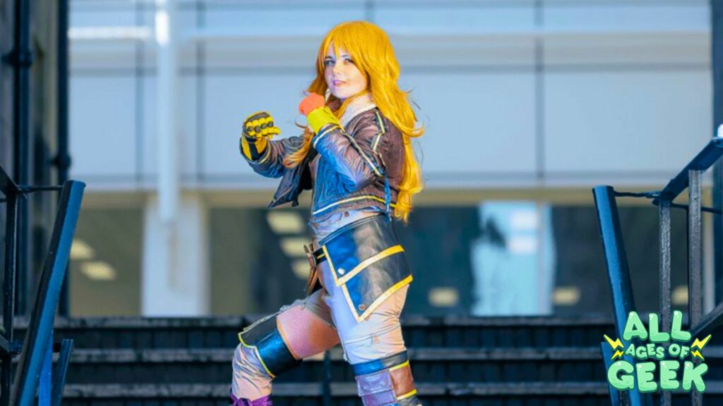 Person cosplaying yang with long yellow hair, wearing a detailed outfit that includes gloves and armor-like accessories, posing confidently on a staircase. The All Ages of Geek logo is visible in the bottom right corner of the image