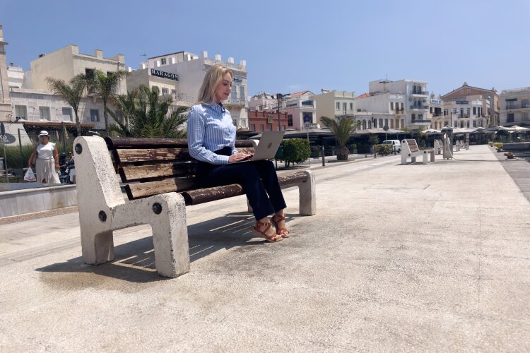 Abby sitting on a bench with her laptop, working while enjoying the beautiful Greek coastal view. 