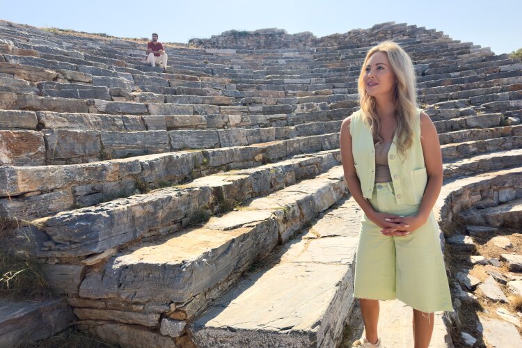 Abby standing in the ancient amphitheater, looking towards Theo who is sitting on the steps.