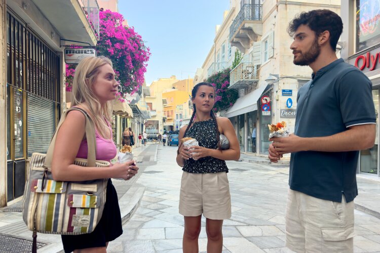Abby, Theo, and a friend enjoying street food in a picturesque Greek village with vibrant flowers in the background.
