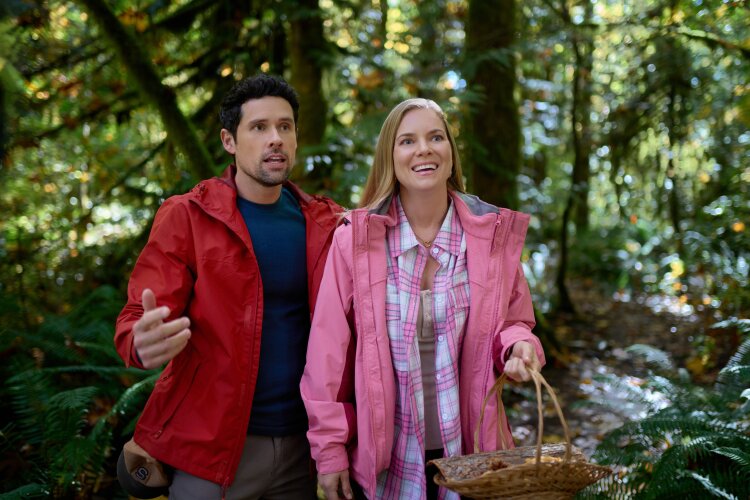 A couple in red and pink jackets standing in a forest, the woman carrying a basket. From Hallmark's "A Whitewater Romance".