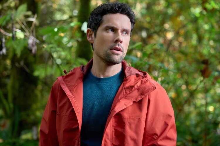  A man in a red jacket standing in a lush forest, looking off to the side with a thoughtful expression. From Hallmark's "A Whitewater Romance".