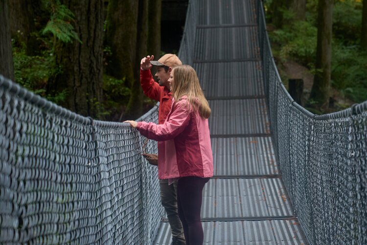 A couple wearing red and pink jackets standing on a suspension bridge, looking out at the scenery in a forest. From Hallmark's "A Whitewater Romance".