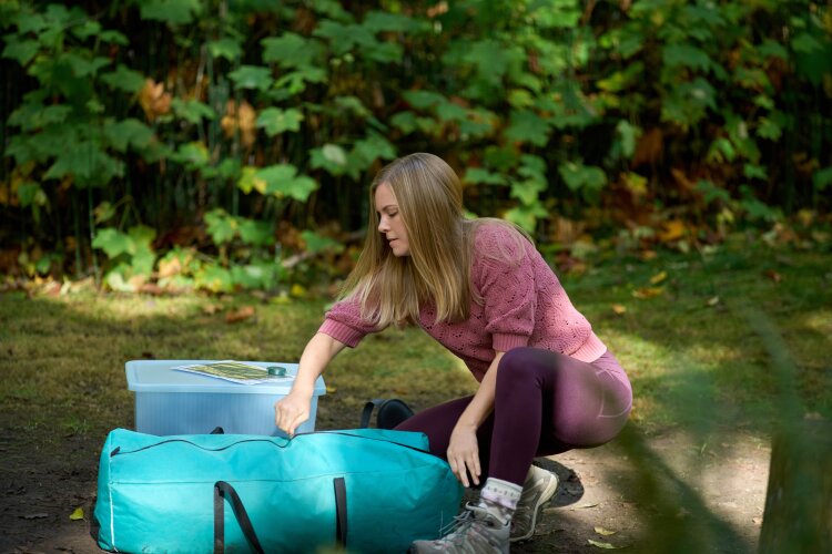 A woman in a pink jacket and purple leggings kneeling on the ground, unpacking a teal duffel bag while camping. From Hallmark's "A Whitewater Romance".