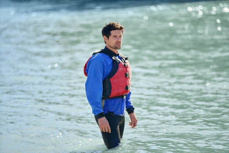 A man standing in shallow water wearing a blue wetsuit and a red life jacket, looking thoughtful. From Hallmark's "A Whitewater Romance".