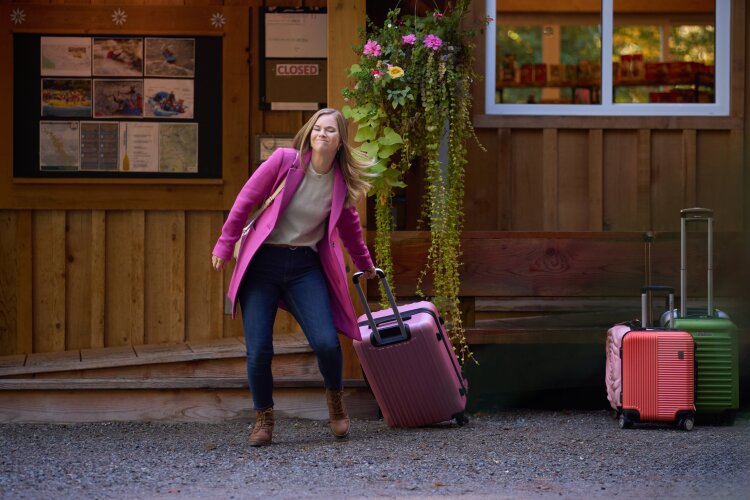 A woman in a pink coat struggling to pull a pink suitcase in front of a rustic lodge. From Hallmark's "A Whitewater Romance".
