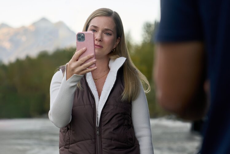A woman holding a smartphone, looking emotional with a backdrop of mountains and trees.