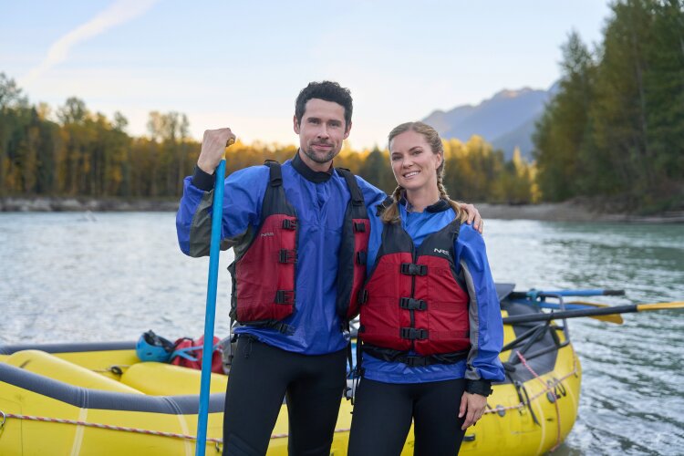 A man and woman in blue wetsuits and red life jackets standing by a yellow raft, smiling at the camera with trees and mountains in the background.