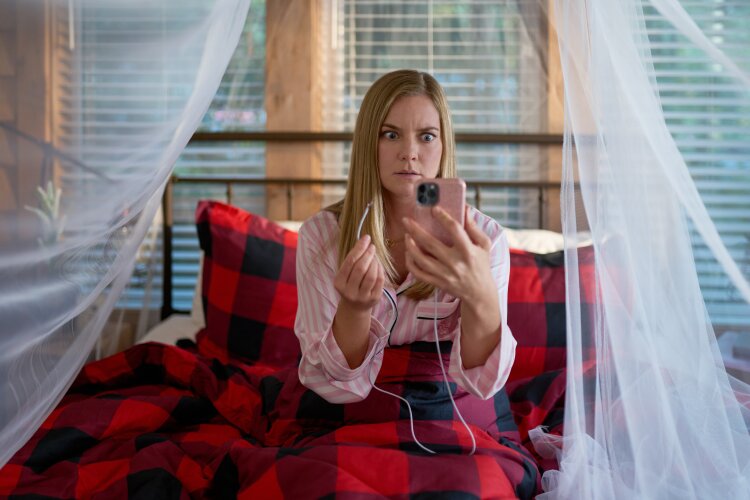 A woman in bed with a surprised expression, holding a smartphone, with plaid bedding and mosquito netting. From Hallmark's "A Whitewater Romance".