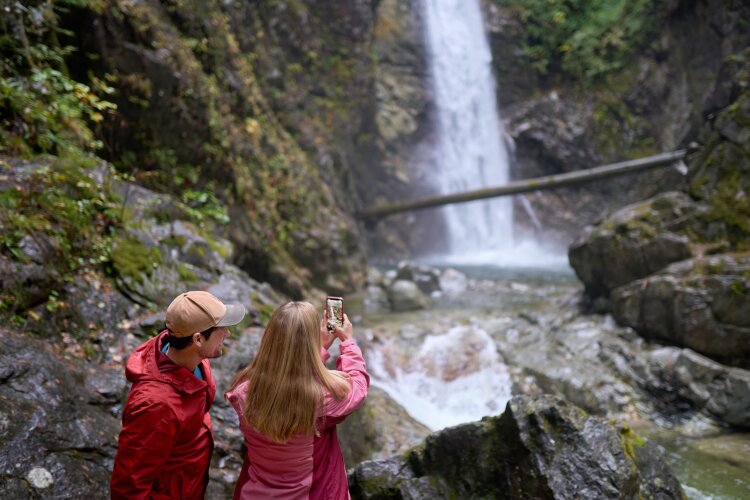  A couple taking a photo of a waterfall with a smartphone, standing on rocks near the water.