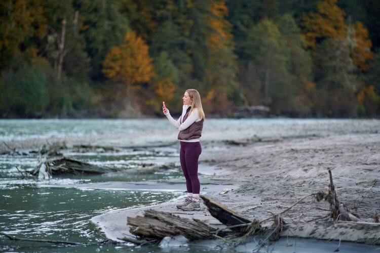 A woman standing on a sandy riverbank, wearing a vest and leggings, taking a photo with her smartphone, with autumn trees and a river in the background. From Hallmark's "A Whitewater Romance".