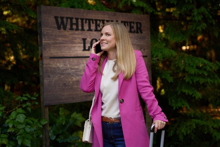 A woman in a pink coat, smiling while talking on her phone and pulling a suitcase, standing in front of a sign that reads "Whitewater Lodge" with greenery in the background. From Hallmark's "A Whitewater Romance".