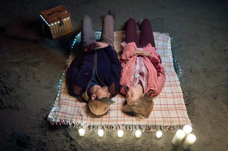 A couple lying on a checkered blanket, surrounded by lit candles and fairy lights, looking at each other lovingly in a cozy setting. From Hallmark's "A Whitewater Romance".