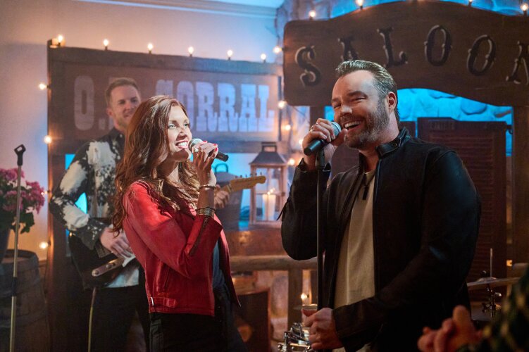  A man and a woman singing into microphones at a lively indoor event, with a band playing in the background. From Hallmark's "Big Sky River: The Bridal Path" ​