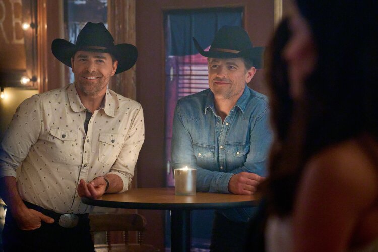 Two men wearing cowboy hats standing inside a dimly lit venue, smiling at someone off-camera. 