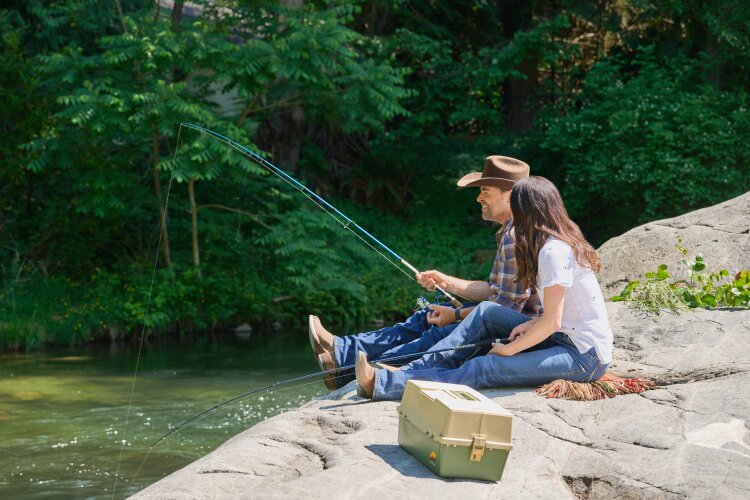 A man and a woman sitting on a rock by a river, fishing and enjoying the scenery. From Hallmark's "Big Sky River: The Bridal Path" ​
