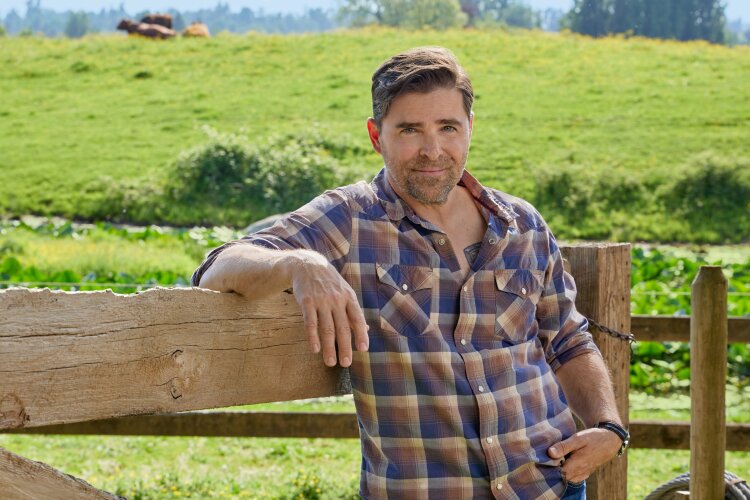 A man in a plaid shirt leaning against a wooden fence, with green fields in the background. From Hallmark's "Big Sky River: The Bridal Path" ​