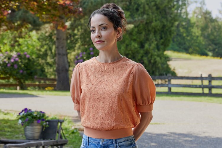 A woman standing outside, wearing an orange top, looking thoughtfully into the distance. 