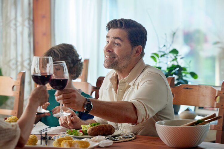 A man sitting at a dinner table, raising a glass of wine with a big smile. From Hallmark's "Big Sky River: The Bridal Path" ​