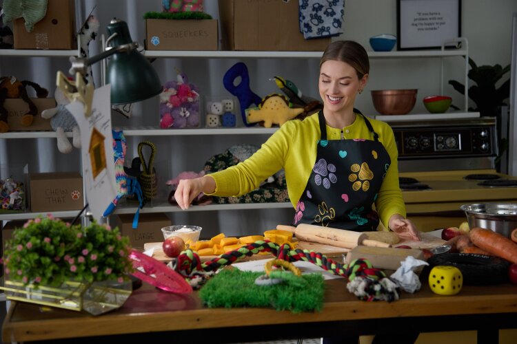  A woman in a brightly colored apron preparing ingredients in a kitchen filled with dog toys and treats.