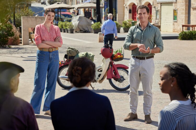 A man and a woman standing in a town square, talking to a small group of people. From Hallmark's "For Love & Honey"
