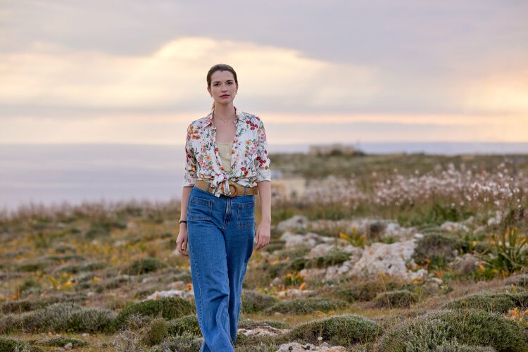 A woman walking in a scenic field at sunset, wearing a floral blouse and blue jeans.
