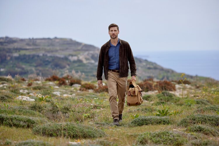 A man walking in a scenic field with a brown leather bag, wearing a blue shirt and brown jacket.