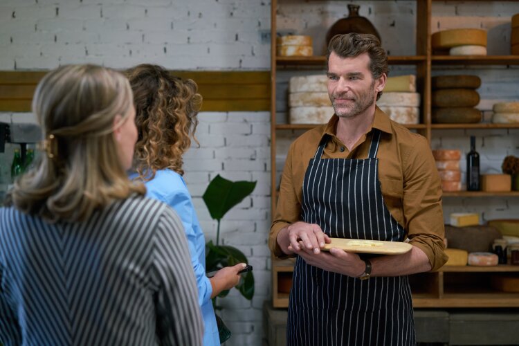 A man in an apron holding a cheese board, talking to two women inside a cheese shop. The shop has a friendly, welcoming vibe with a rustic décor.