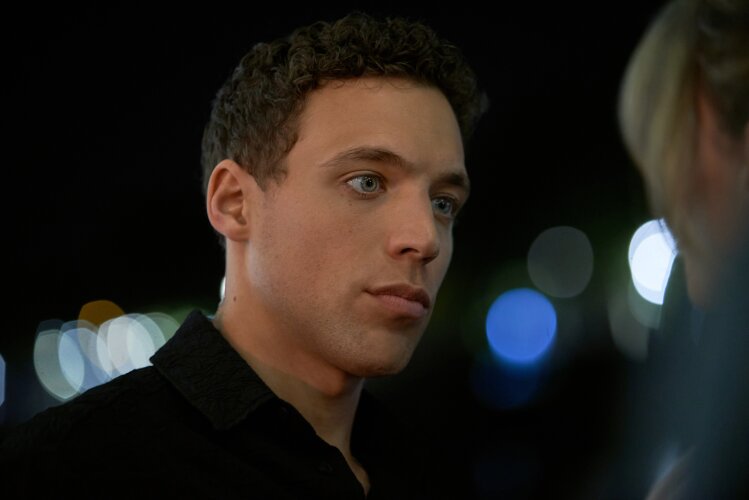 A close-up of a man with short curly hair, looking intently at someone off-camera, with blurred lights in the background.