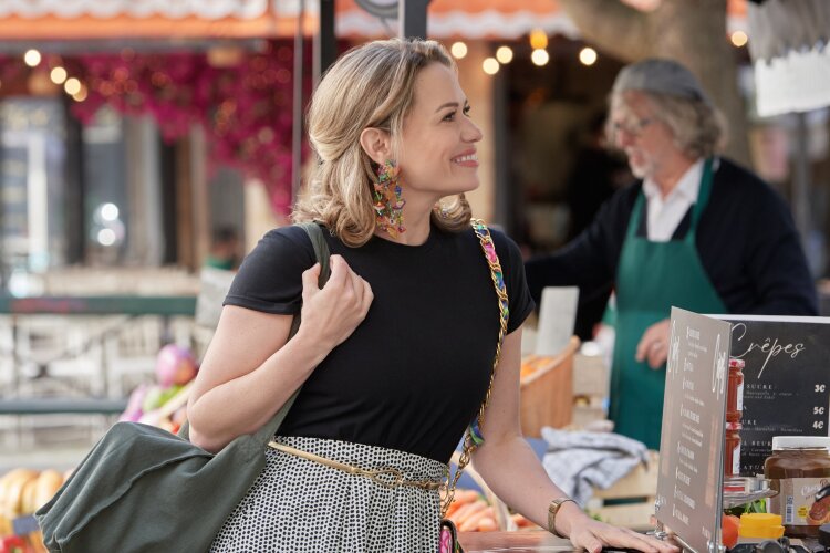 A woman shopping at an outdoor market, smiling and carrying a bag, with colorful produce and market stalls around her.