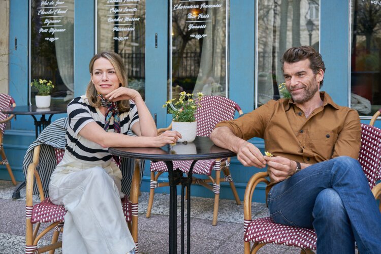 A man and a woman sitting outside a café, smiling and talking while enjoying their drinks. The café has a charming, classic Parisian style with small tables and checkered chairs.