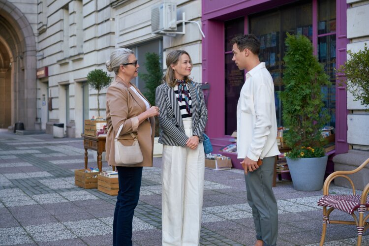 Three people standing and conversing on a sidewalk outside a shop. The woman in the center is engaged in conversation with an older woman and a younger man.